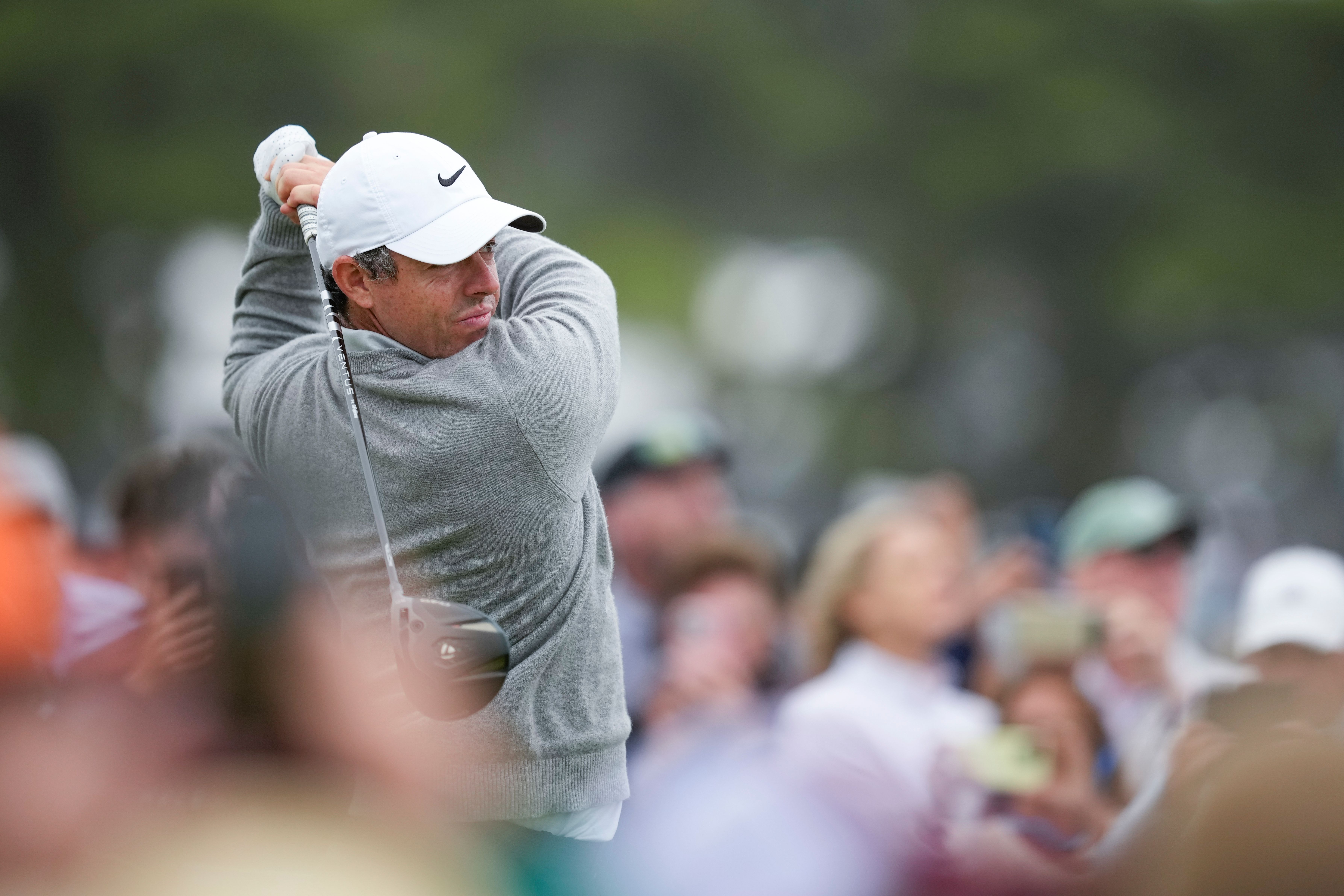 A male golfer in a white cap and grey shirt powerfully swings a golf club during a tournament.
