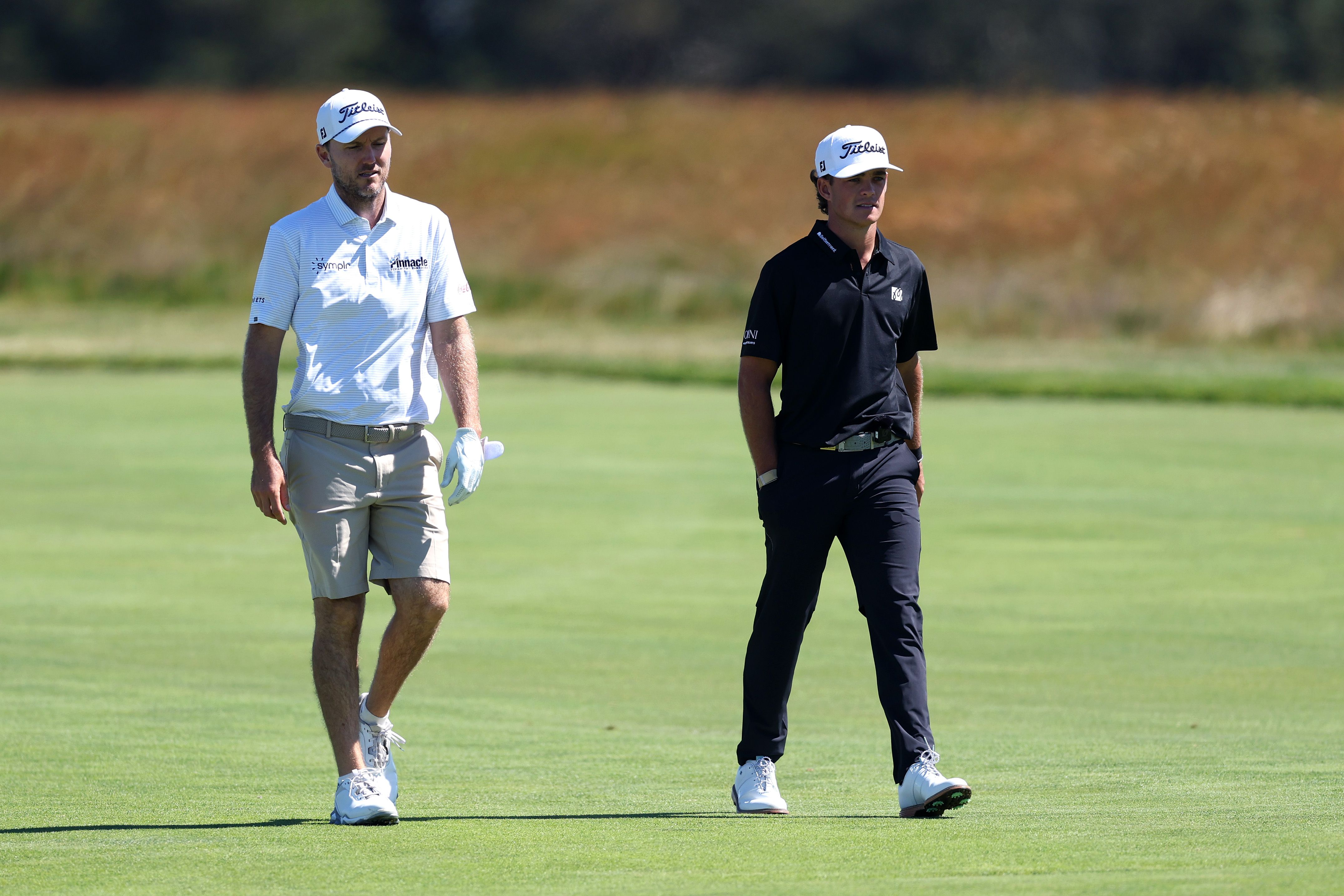 Two male golfers in caps and polos walk on a bright green golf course.