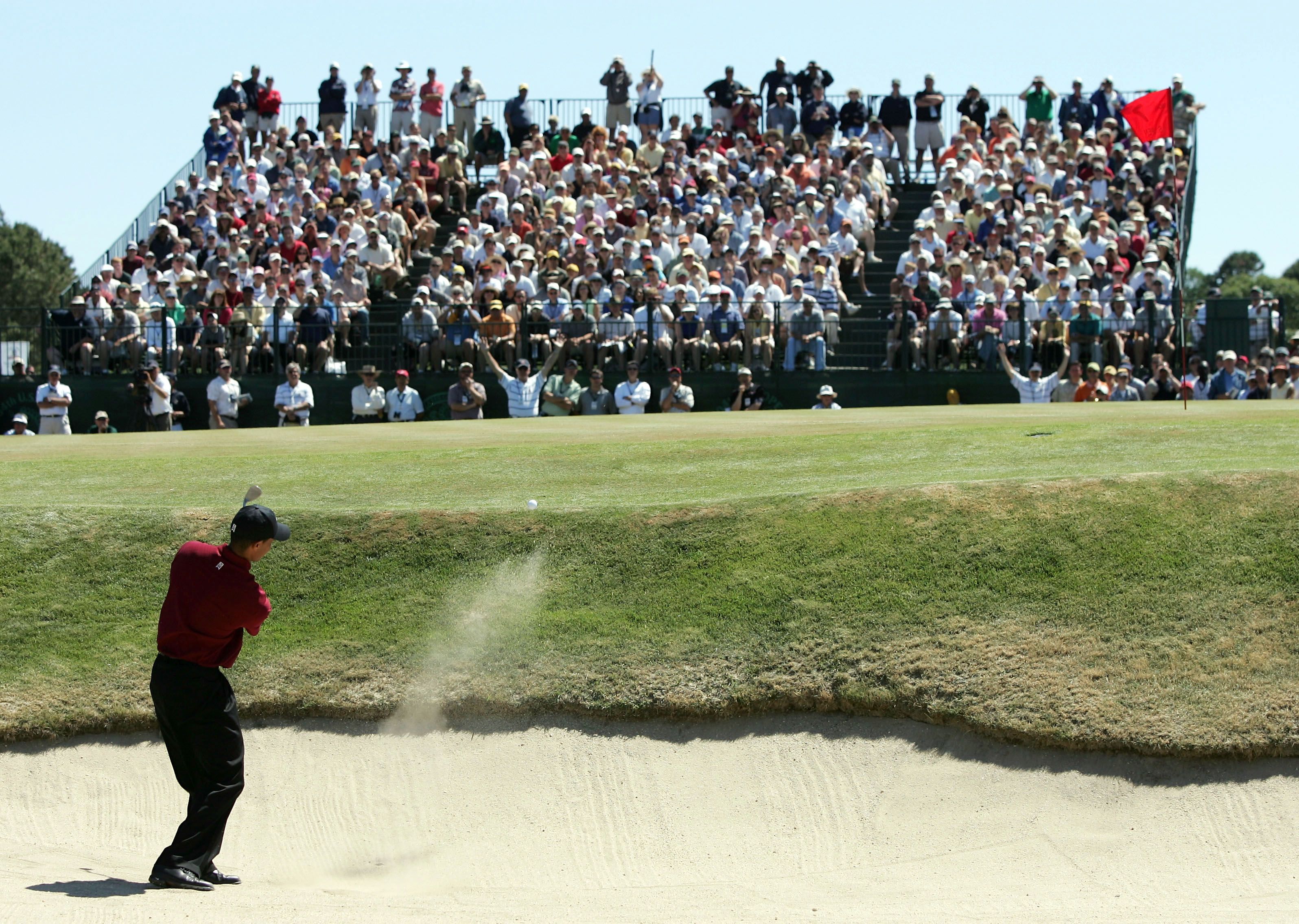 Golfer in red shirt hits ball from sand bunker with crowd watching.