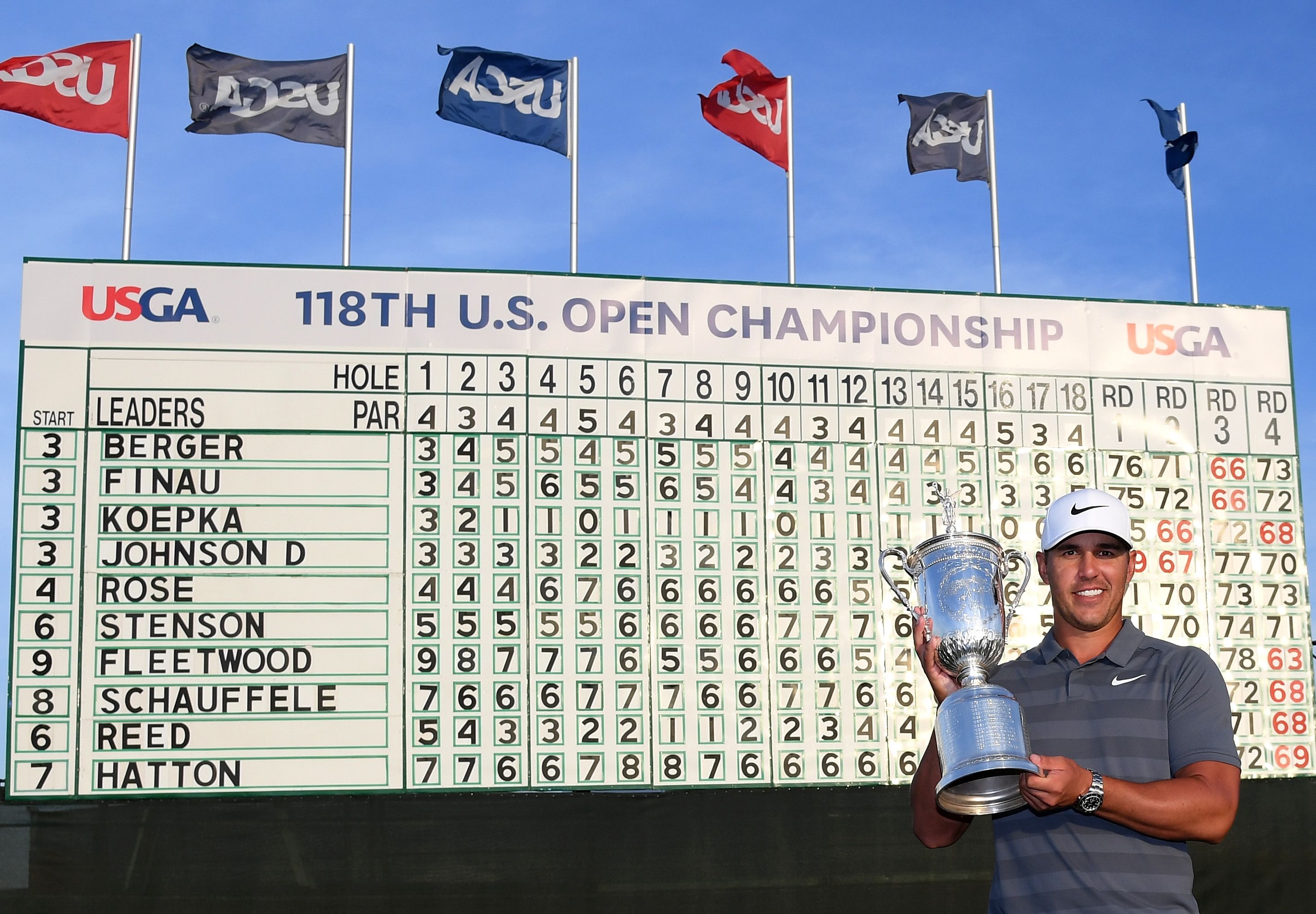 Golfer Brooks Koepka holds the U.S. Open trophy beside a large championship scoreboard.