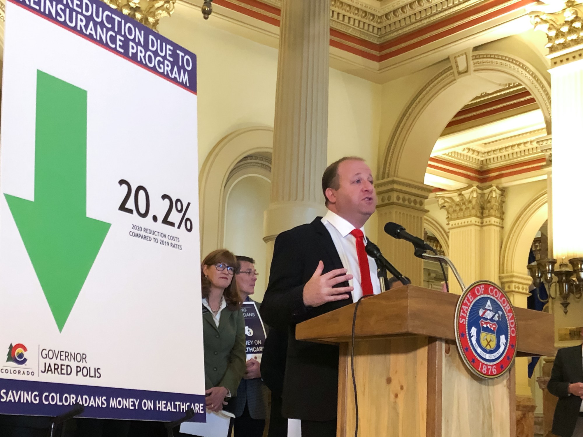 Gov. Jared Polis speaking at a podium in a government building, next to a visual display showing a 20.2% reduction due to the reinsurance program for Colorado's healthcare costs.