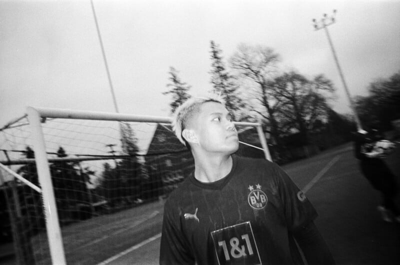 A person in a Borussia Dortmund soccer jersey stands in front of a goal on an outdoor field, looking to the side. The image is in black and white, with trees and light poles visible in the background.