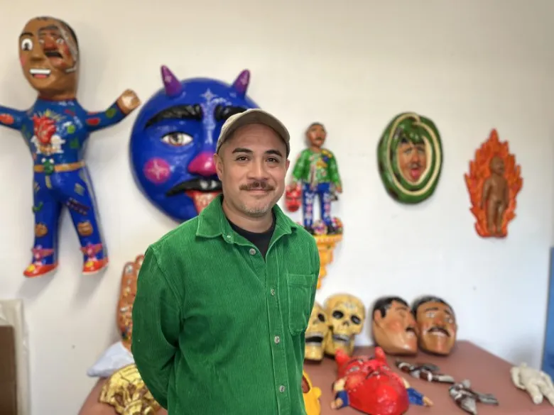 A man in a green shirt stands in front of a wall displaying colorful masks and figurines.