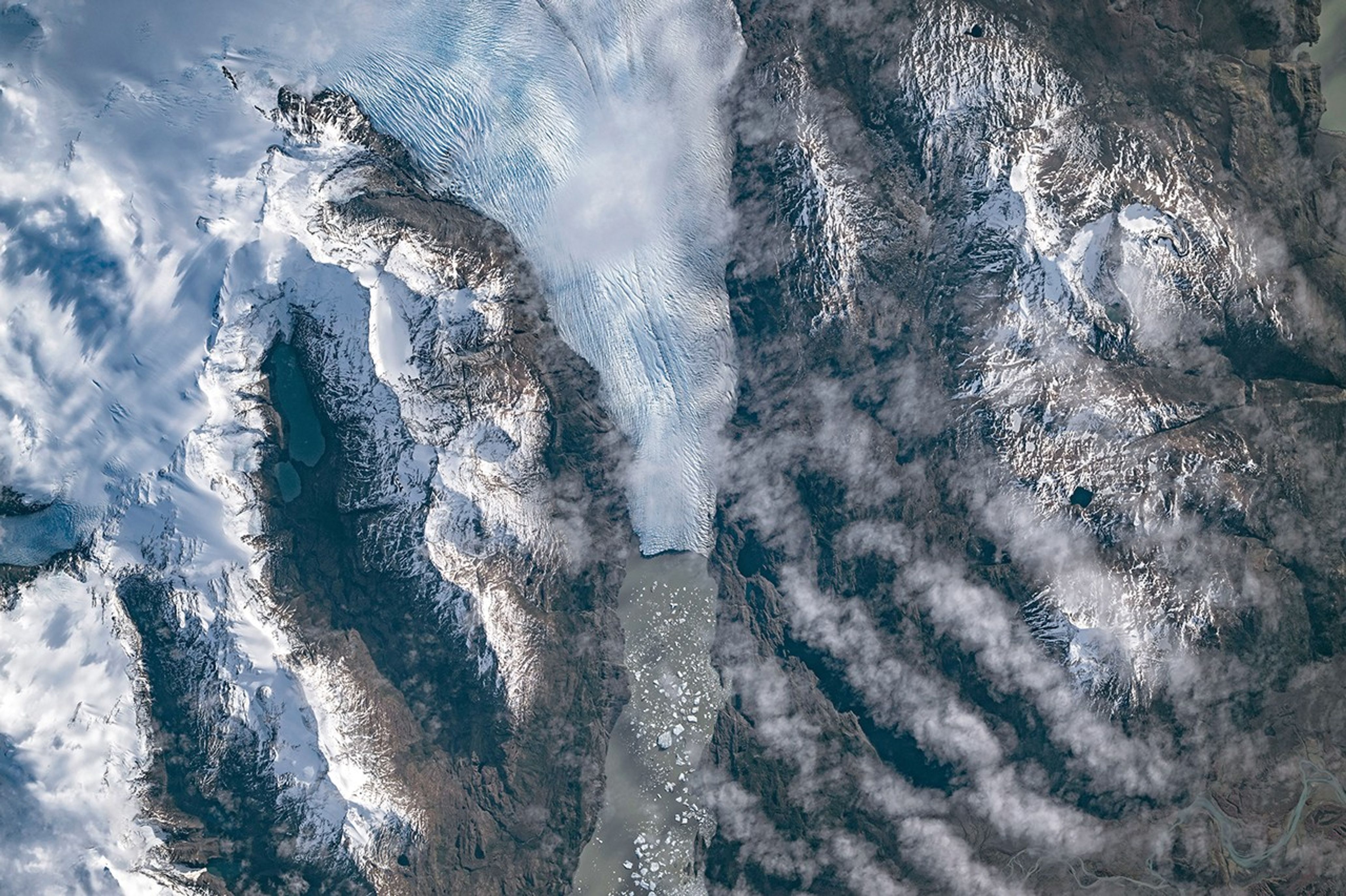 A top-down photo shows a glacier running from north to south amid snowy, mountainous terrain. The glacier flows into a narrow lake where many small icebergs are visible floating on the water.