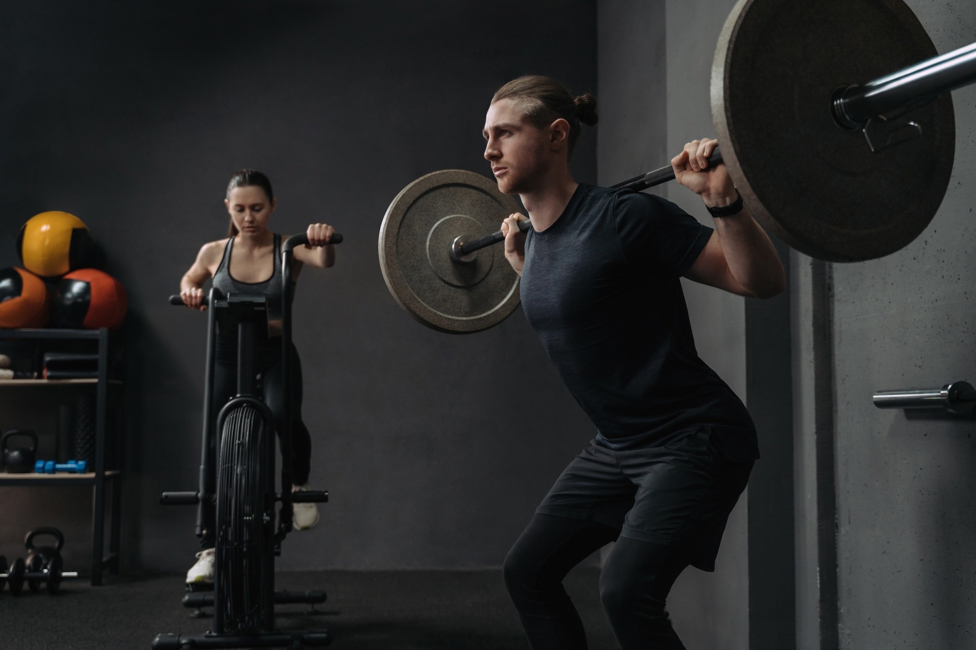 Fit woman is working out on exercise bike, while muscular man training with barbell.