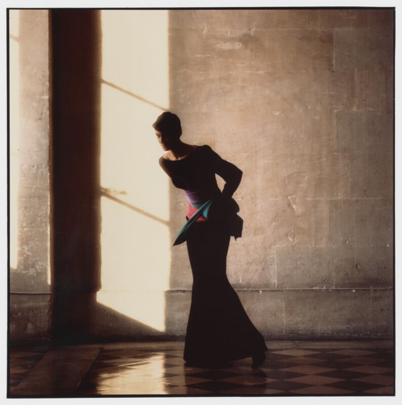 A fashionably dressed woman stands in profile against a sunlit wall, casting a shadow. She wears a long, dark gown with colorful accents and poses gracefully on a checkered floor.