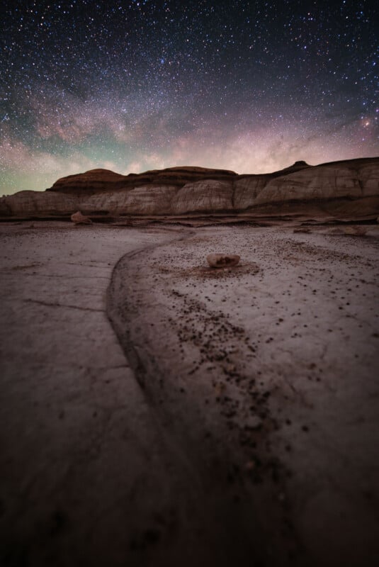 A rocky desert landscape under a clear, star-filled night sky. The foreground shows textured, pale rock with scattered stones, while layered rock formations stretch across the horizon.