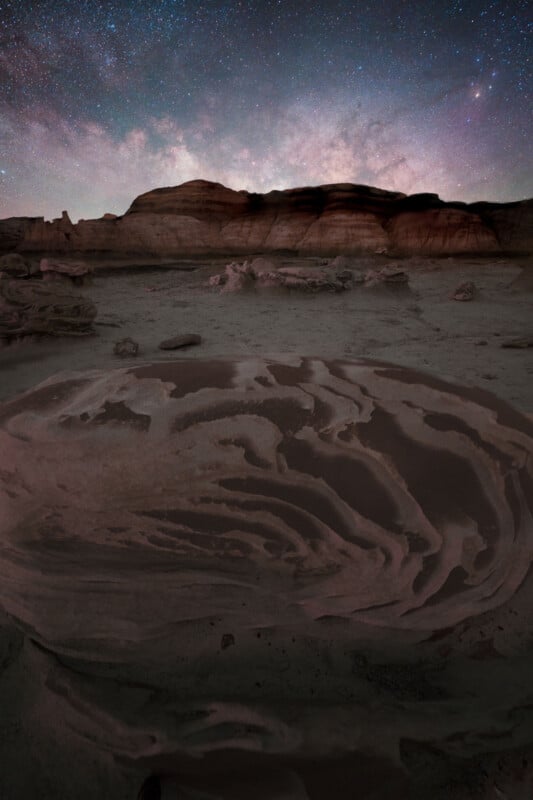 A surreal desert landscape features swirling rock patterns in the foreground, rugged cliffs on the horizon, and a star-filled night sky with the Milky Way arching above.