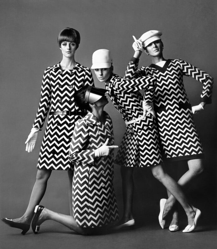 Four women pose in matching zigzag-patterned dresses and hats, striking playful, dramatic poses against a plain background in a black and white fashion photo.