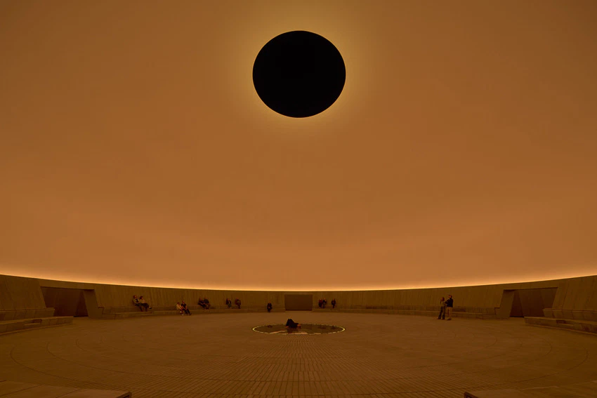 Skyspace installation with glowing orange interior and circular black opening above. People seated around the space's perimeter.