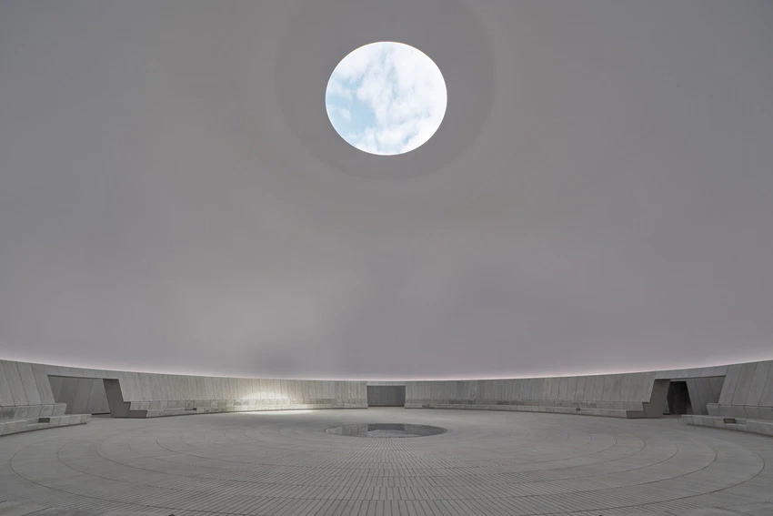 Modern circular concrete structure with a large circular opening in the ceiling, showcasing blue sky and clouds above.