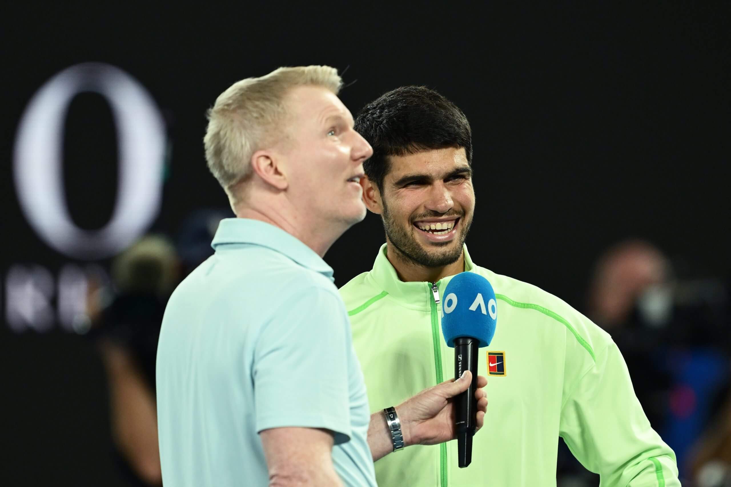 Jim Courier looks up holding a blue AO-branded microphone as Carlos Alcaraz stands next to him wearing a light green Nike jacket.