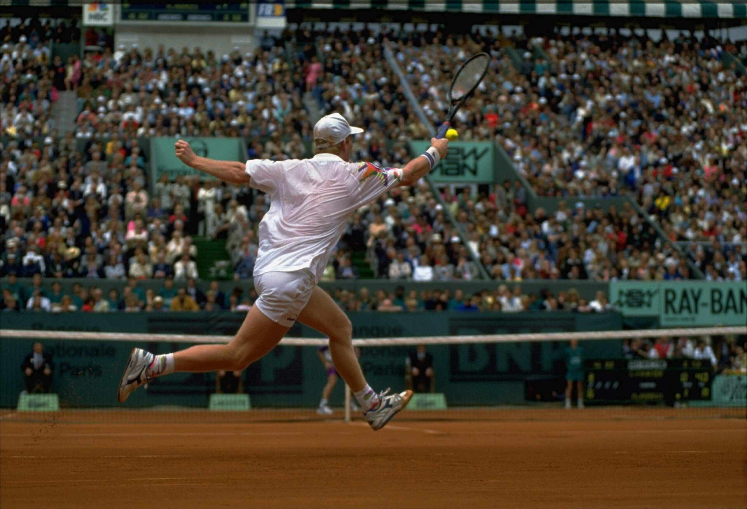 A shot of Jim Courier from behind as he hits a forehand on the run on a clay court, with the camera at court level.