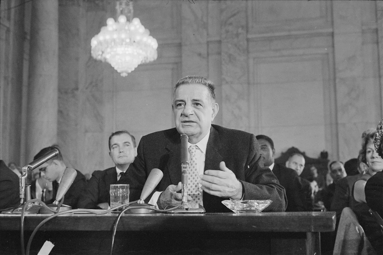 In this black-and-white photo, Valachi sits in a congressional chamber. On the table in front of him are several vintage microphones, a glass of water, and a glass ashtray. A group of people sits behind him listening attentively. The setting is a grand hall or hearing room featuring marble columns and a large, ornate crystal chandelier hanging illuminated from the ceiling.