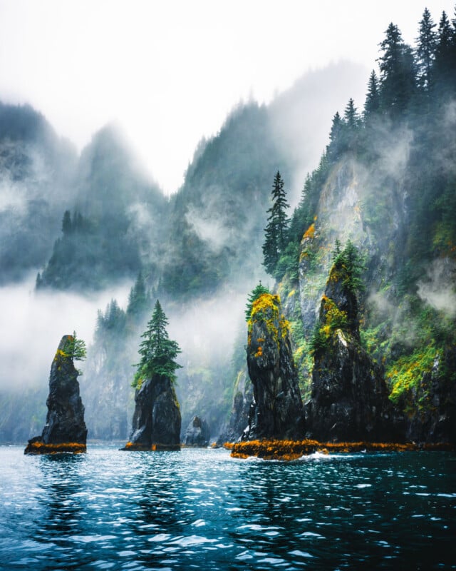 Rocky sea stacks topped with green trees rise from blue water, with mist and fog drifting among tall, forested mountains in the background, creating a dramatic and serene landscape scene.