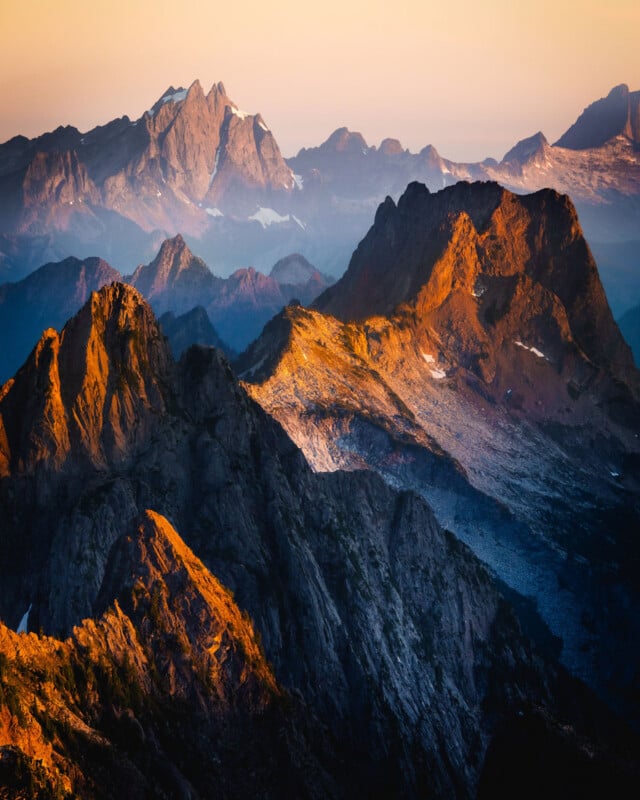 Mountain range at sunrise with rugged peaks and ridges bathed in warm golden light, casting dramatic shadows. Distant mountains fade into the soft hues of morning, creating a layered landscape under a clear sky.