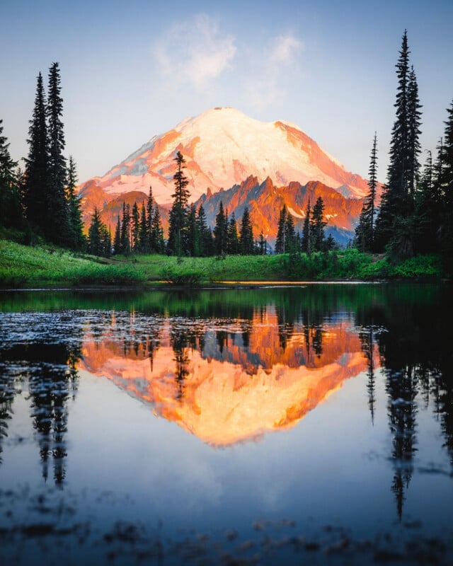 Snow-capped mountain at sunrise or sunset, glowing with orange light, reflected in a calm lake surrounded by evergreen trees and green grass under a clear sky.