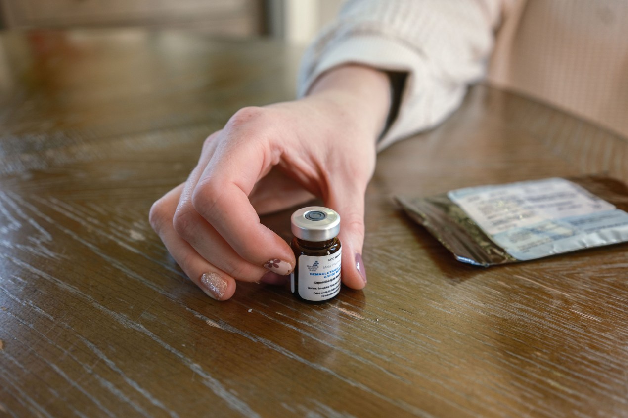 A woman's hand holds a small vial of liquid GLP-1 medication on a table.
