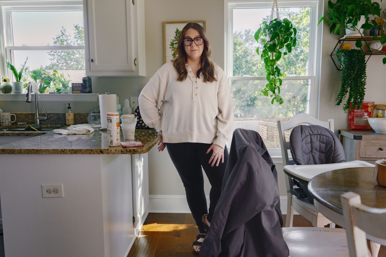 A woman stands beside her kitchen counter and dining table and faces the camera.