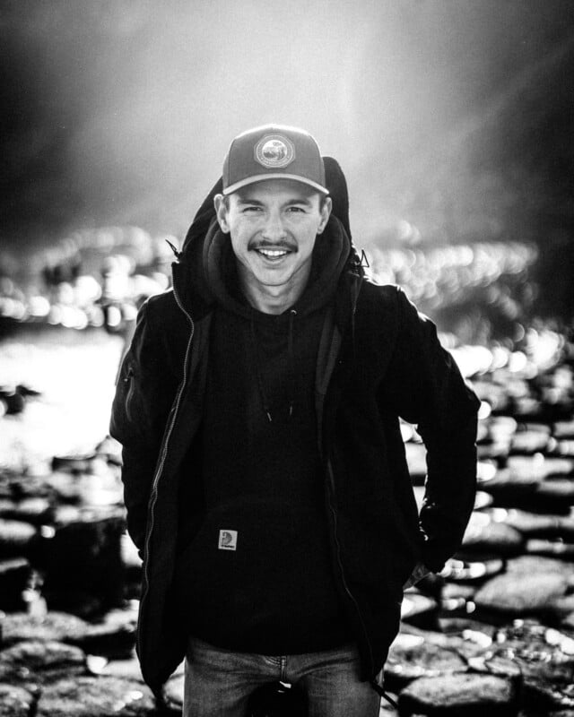 A man wearing a cap and jacket stands outdoors, smiling at the camera with hands in his pockets. The background is rocky and sunlit, creating a dramatic light effect. The photo is in black and white.