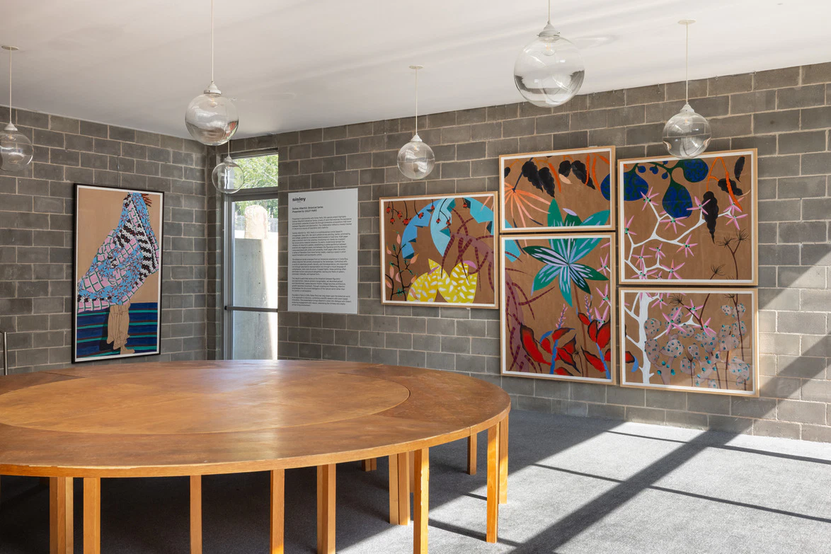 Art exhibit in a modern gallery showcasing colorful abstract paintings on gray brick walls with a round wooden table in the center.