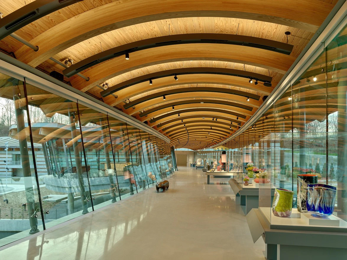 Modern art gallery with wooden arched ceiling, large windows, and vibrant glass sculptures displayed on the right.