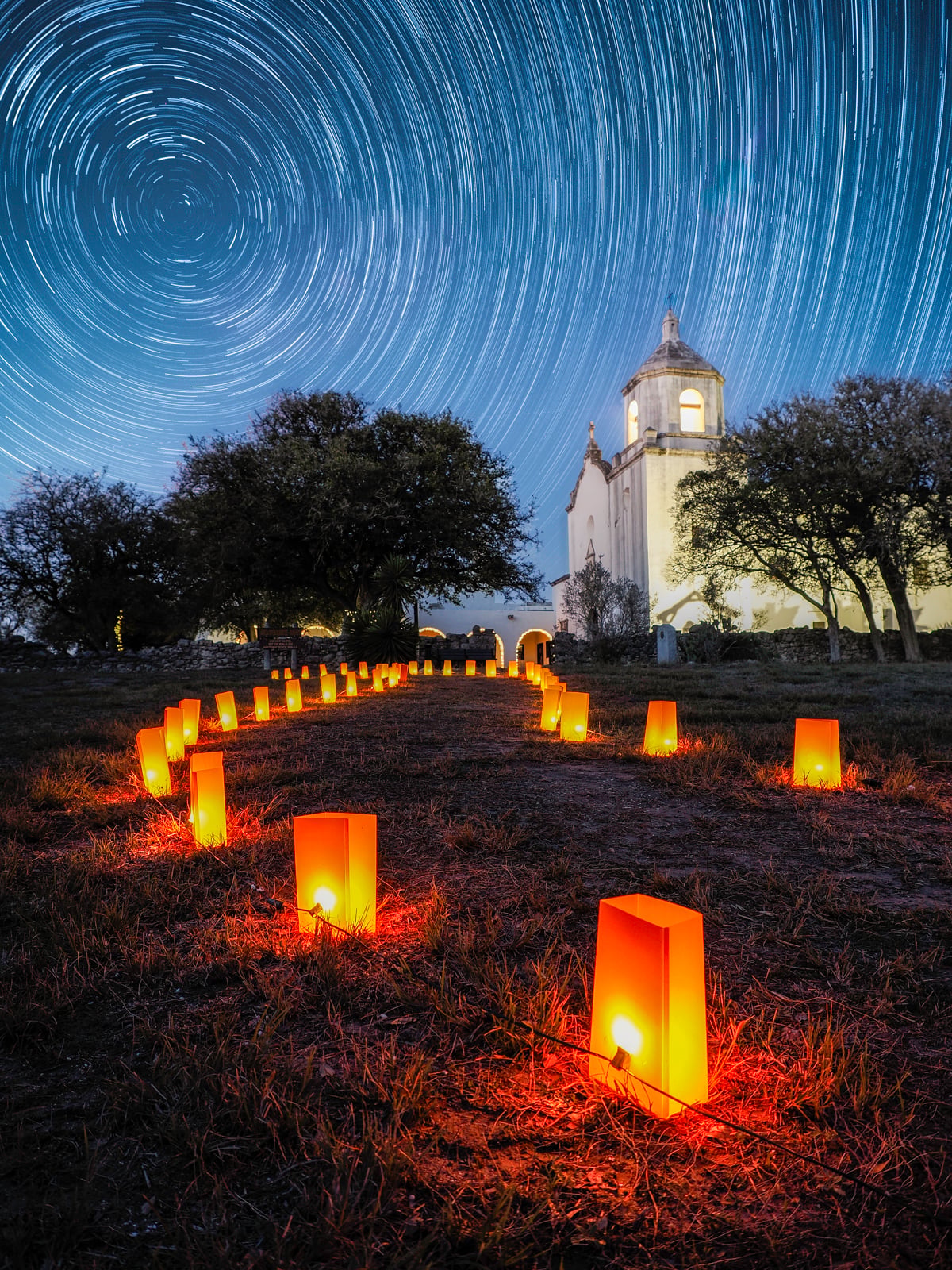 A path lined with glowing lanterns leads to a white church at night, surrounded by trees. Above, star trails create circular patterns in the dark sky, emphasizing the passage of time.