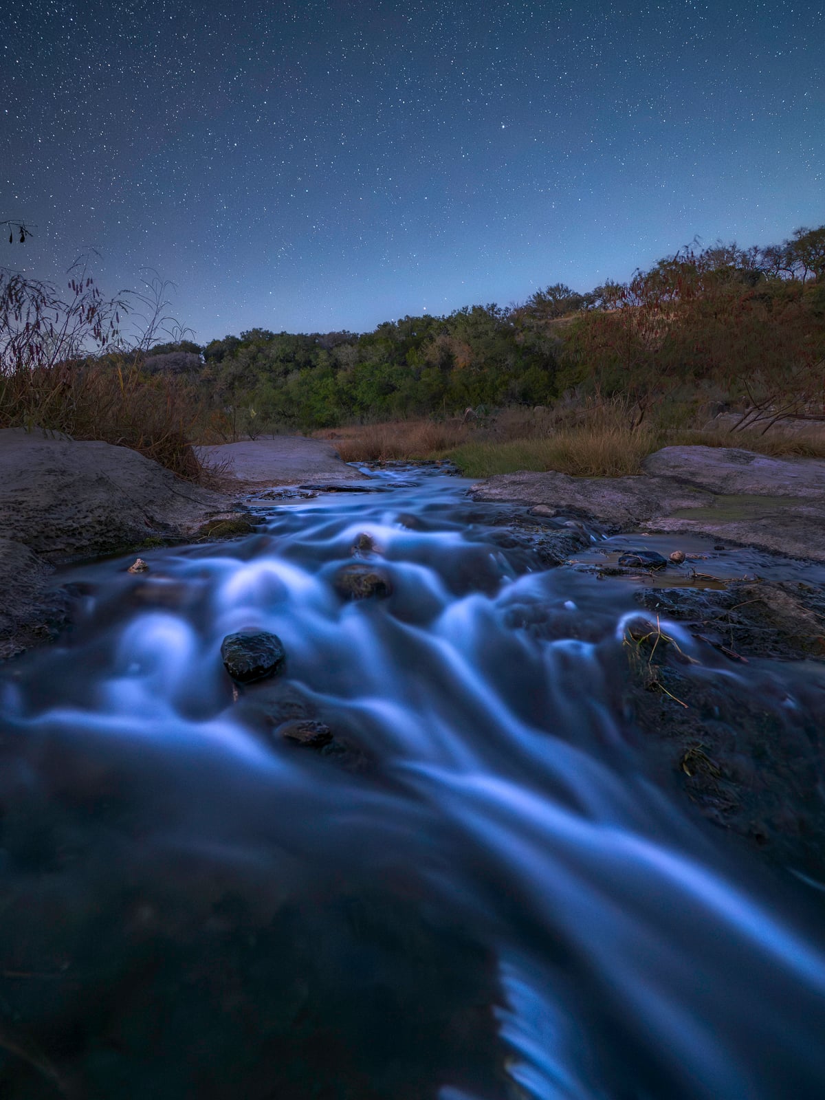 A small, smooth-flowing stream cascades over rocks under a starry night sky, surrounded by grass and trees, creating a tranquil, long-exposure effect in the natural landscape.