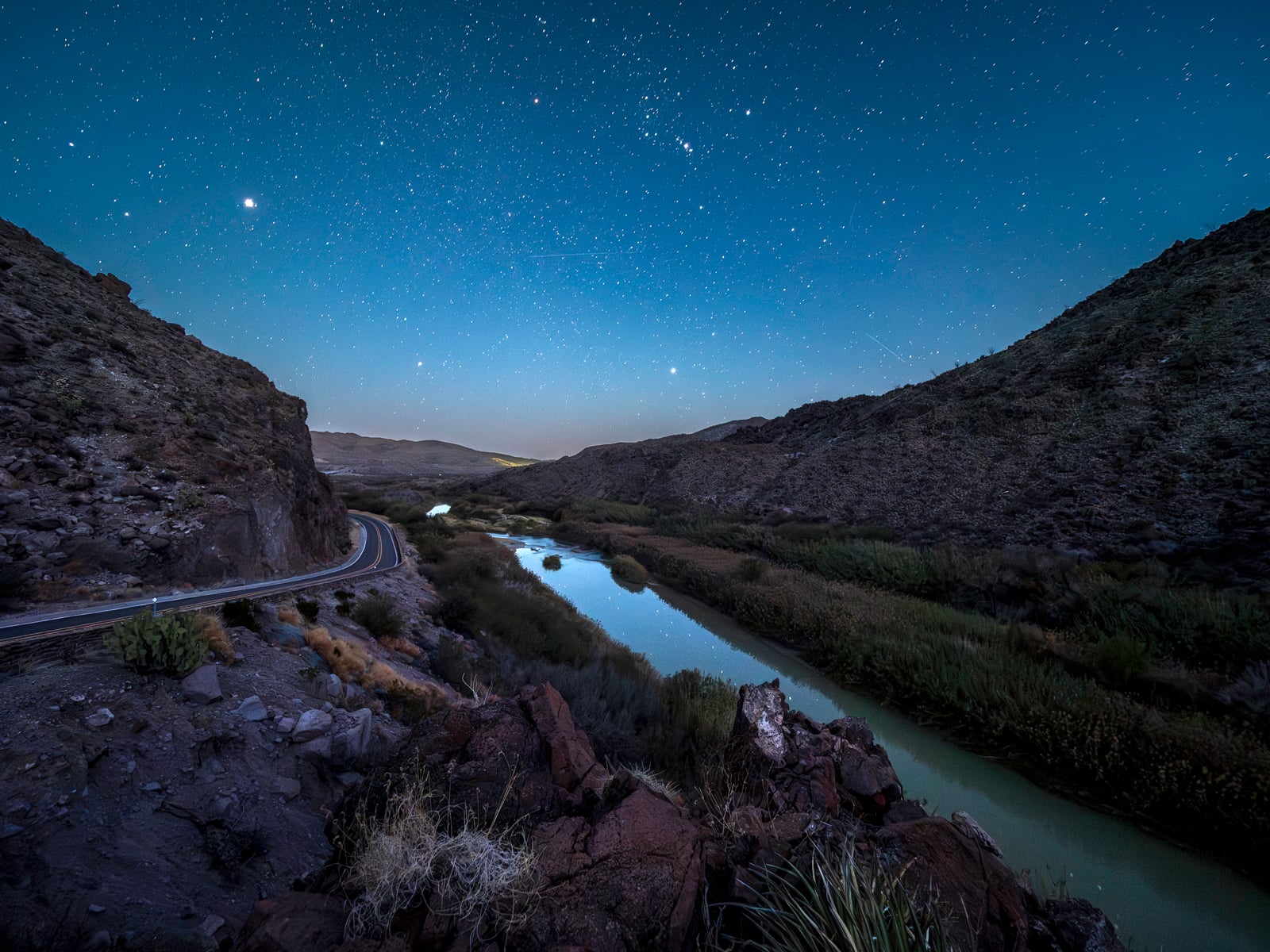 A winding road runs alongside a river between rocky hills under a clear, starry night sky, with reflections of stars visible in the calm water.