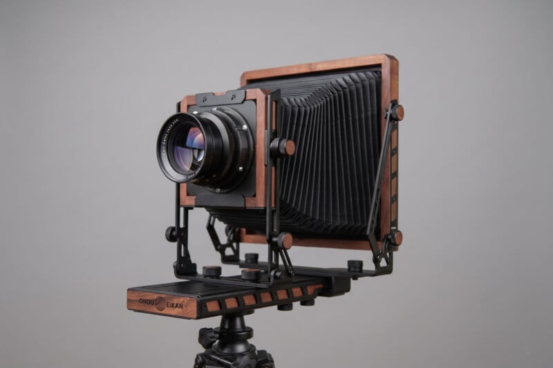 A large format wooden field camera with a black bellows, mounted on a tripod, is shown against a plain gray background. The camera features metal and wood components, giving it a classic, vintage look.