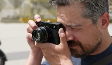 A man with short hair and a beard is holding a black camera up to his face, looking through the viewfinder as he takes a photo outdoors in bright daylight.