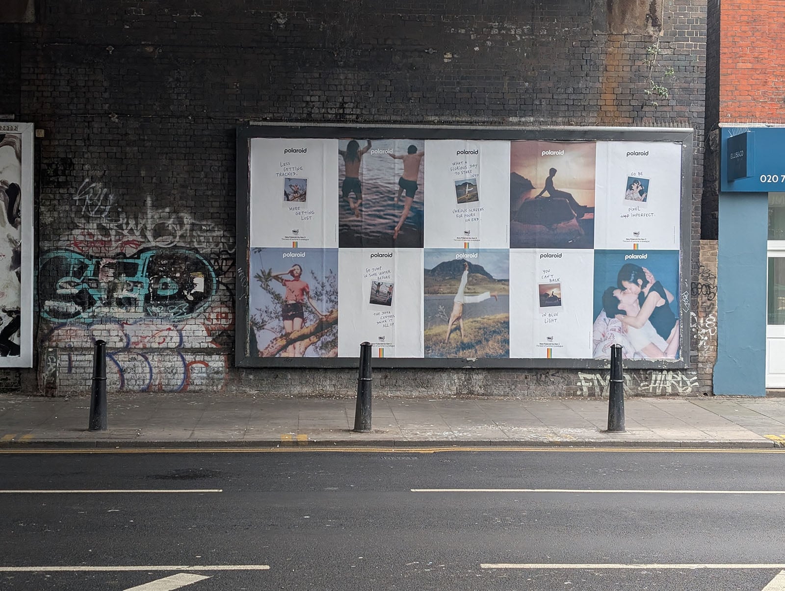 A street scene shows a large billboard with Polaroid-themed posters, featuring various photos of people and nature, mounted on a brick wall with graffiti. Three black bollards line the sidewalk in front.