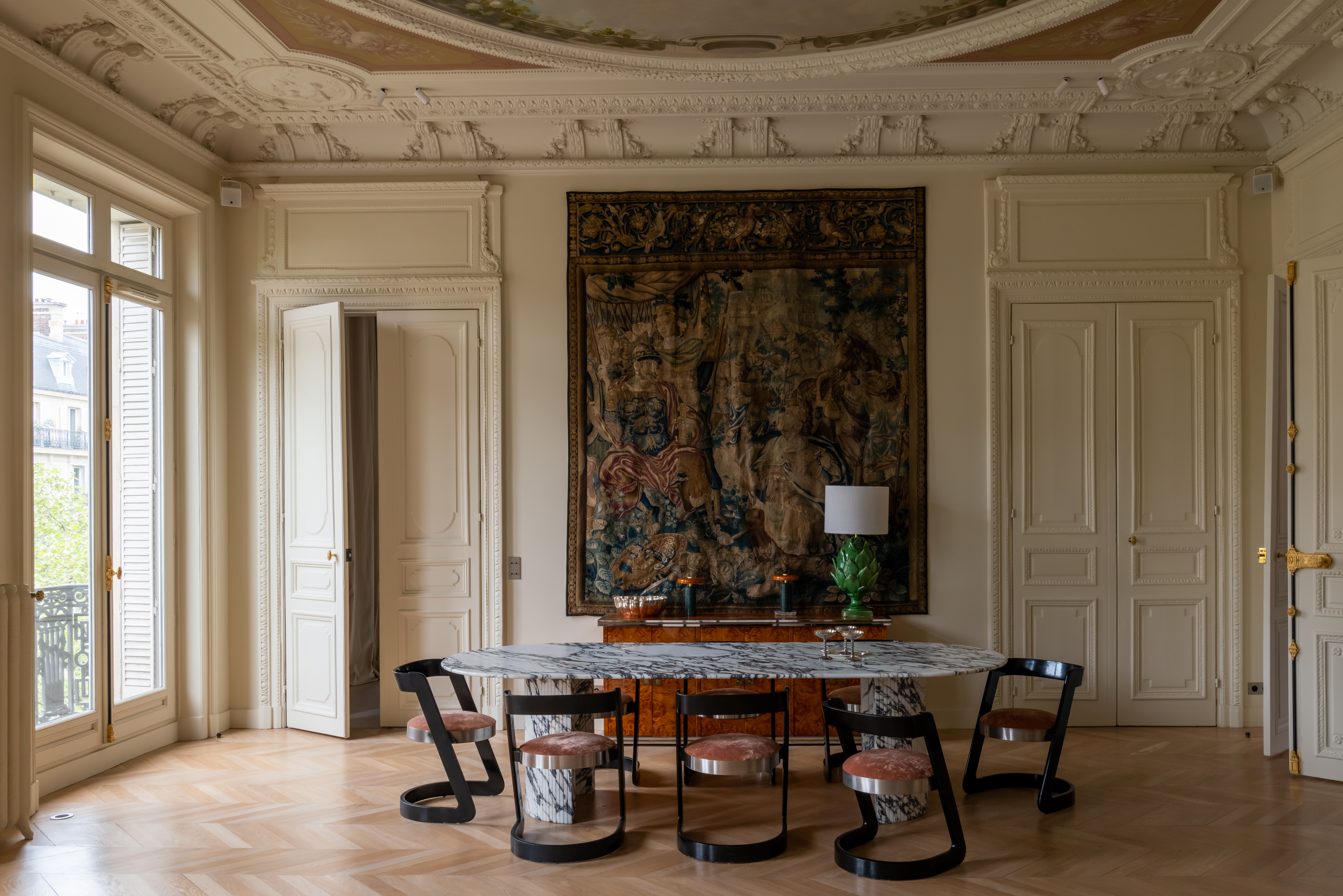 A grand dining room with a marble oval table and statement dining chairs with black backs and legs, and round seating with a large, dark tapestry hanging on an off-white white wall and an open door