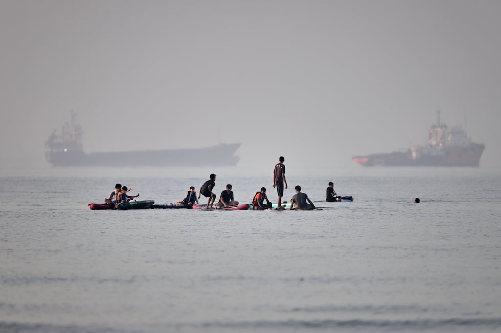 People gather on paddleboards in shallow water as cargo and service vessels are anchored in...