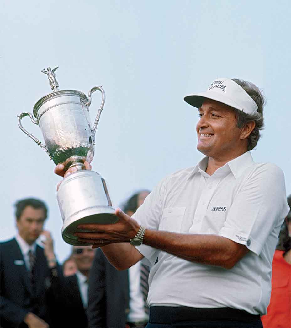 A man in a white shirt and visor smiles while holding up a large silver trophy with both hands, celebrating his U.S. Open win at Shinnecock Hills. Several people stand in the background beneath the clear sky.