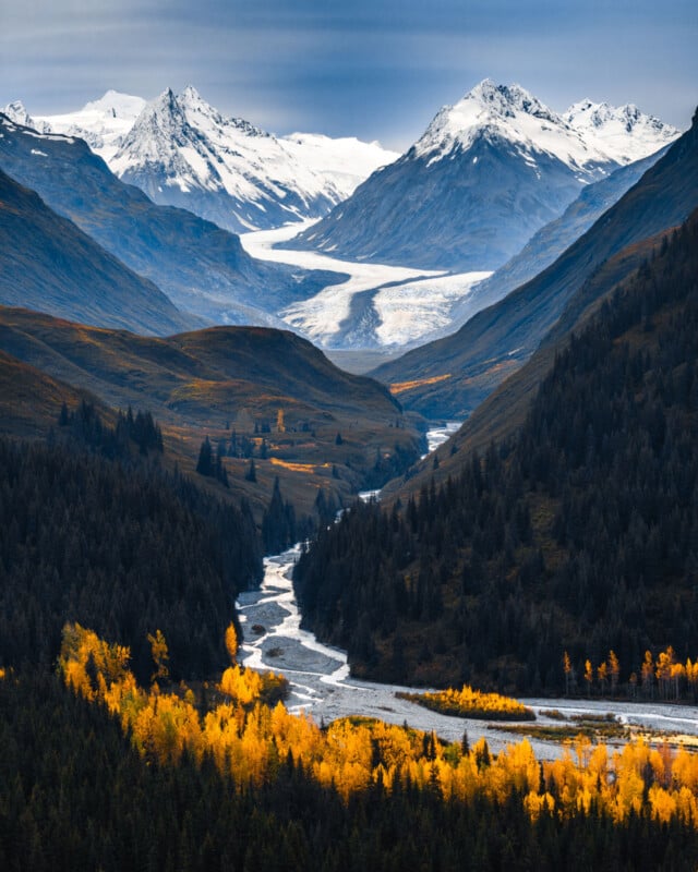 A winding river flows through a forested valley with bright yellow autumn trees, leading toward snow-capped mountains and a glacier in the distance under a cloudy sky.