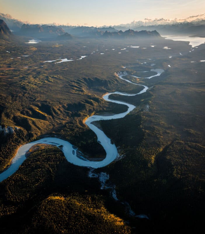 A winding river snakes through a vast, forested landscape under golden sunlight, with distant mountains and lakes visible on the horizon.