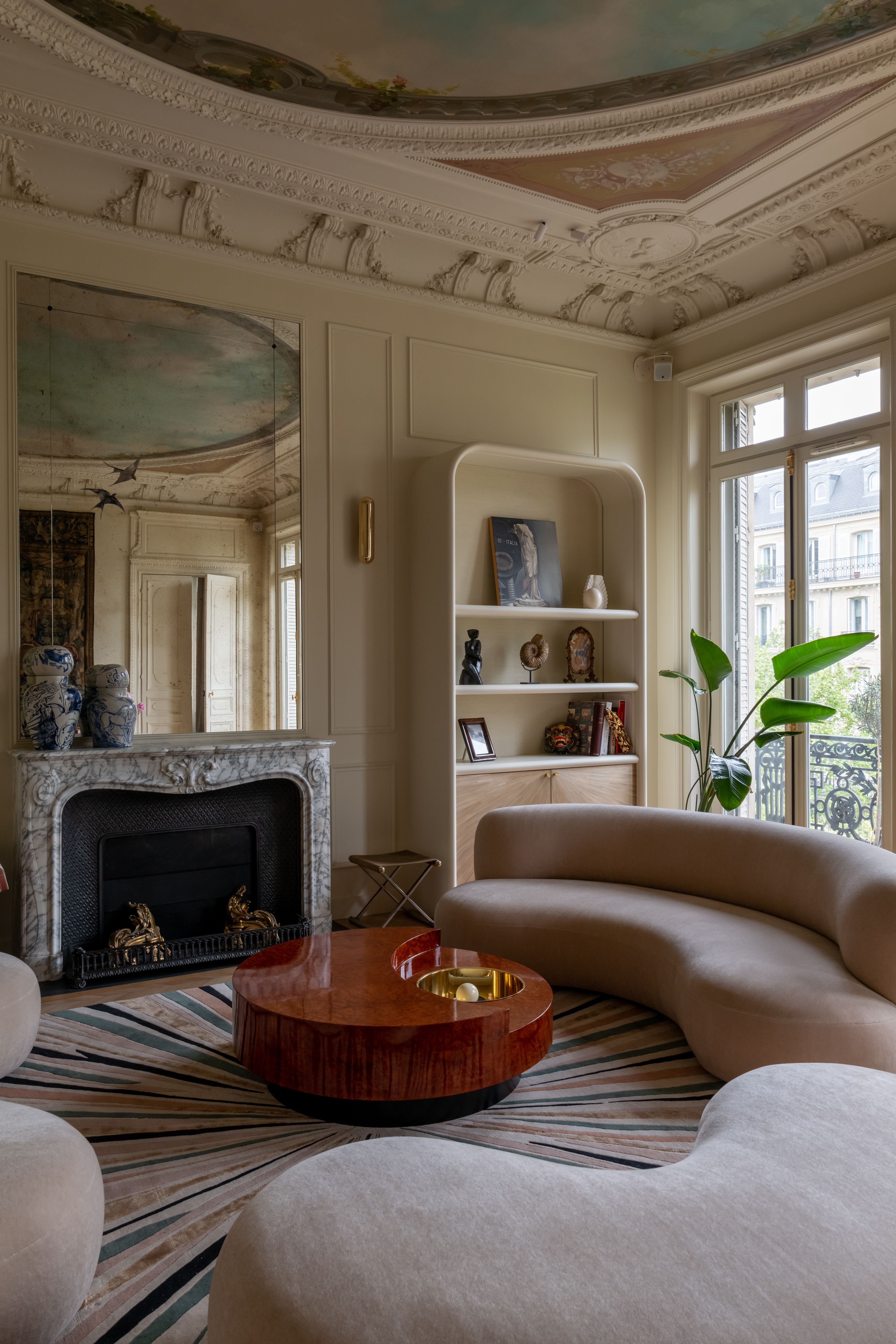 A contemporary living room with bluh pink curved sofas, and elaborate circular painted ceiling, and a large mirror above the fireplace with a bookcase and plant by the window