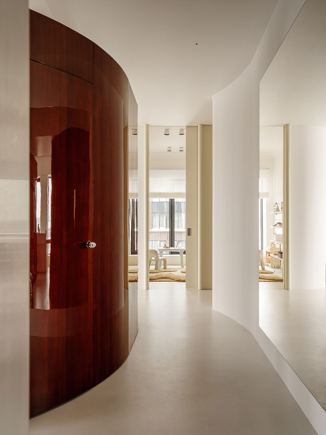 A modern hallway with curved white walls, a large mirror, and a glossy Antwe wooden door leads to a bright living area with floor-to-ceiling windows.