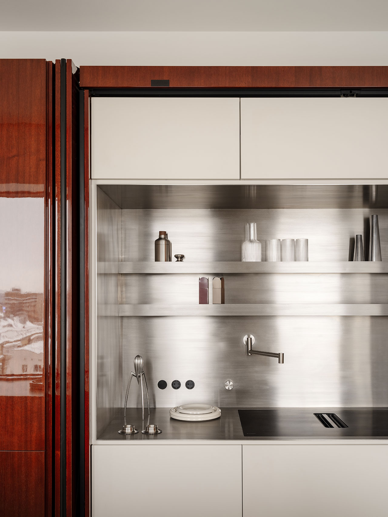 Minimalist Antwe kitchen with glossy wood panels, stainless steel backsplash, built-in sink, stovetop, shelves with containers, and a view of buildings outside.