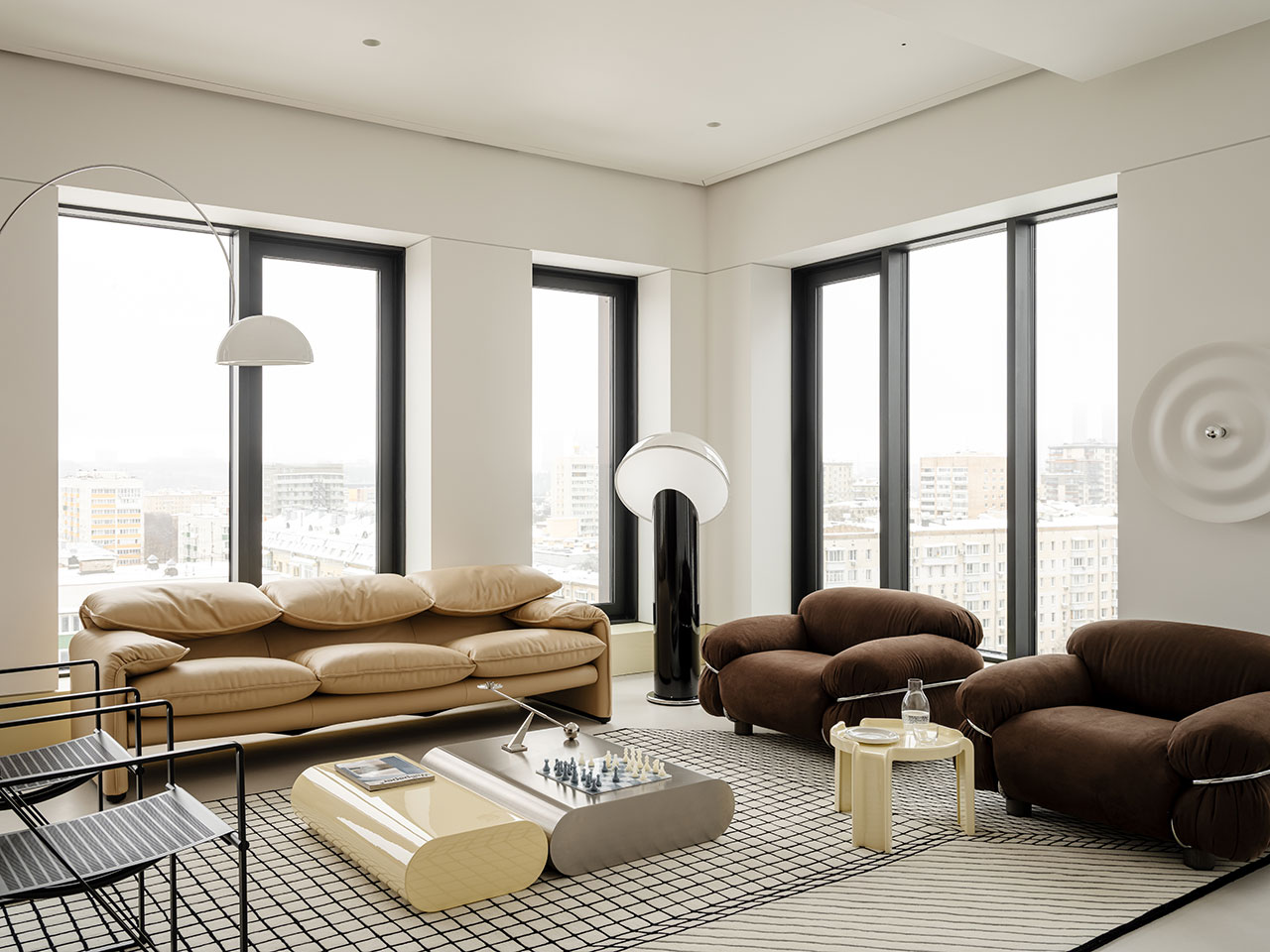 Modern Antwe living room with large windows, beige and brown sofas, geometric coffee tables, a chessboard, and contemporary floor lamps overlooking a cityscape.