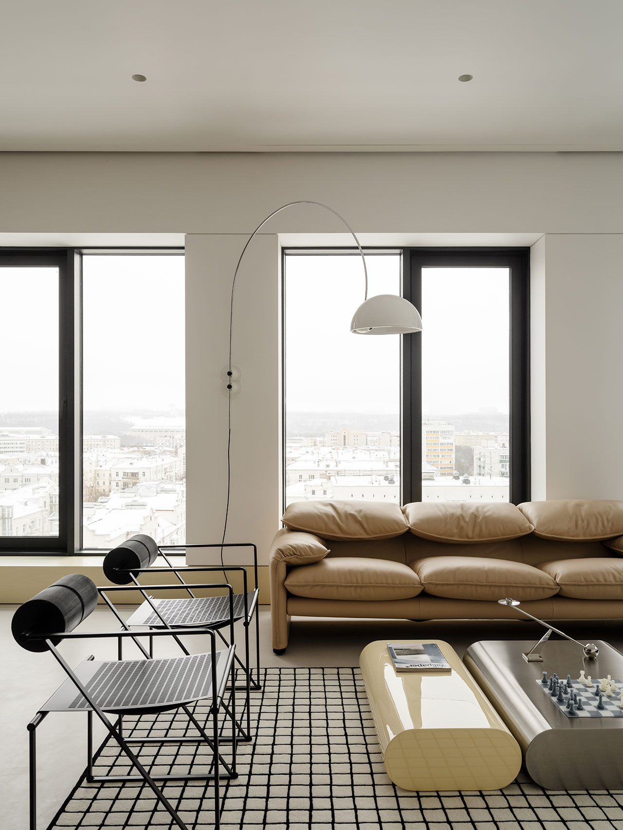 Modern Antwe living room with large windows, beige sofa, minimalist chairs, a grid-pattern rug, sleek coffee table, and a curved floor lamp, overlooking a stunning cityscape.