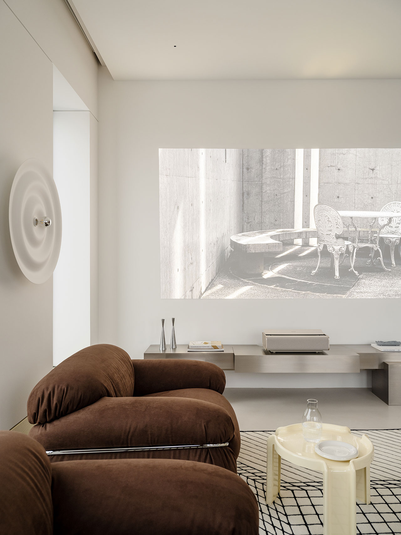 Modern living room with Antwe brown lounge chairs, a low beige table with glassware, and a wall projection of a black-and-white outdoor scene. Minimalist decor with neutral tones completes the look.