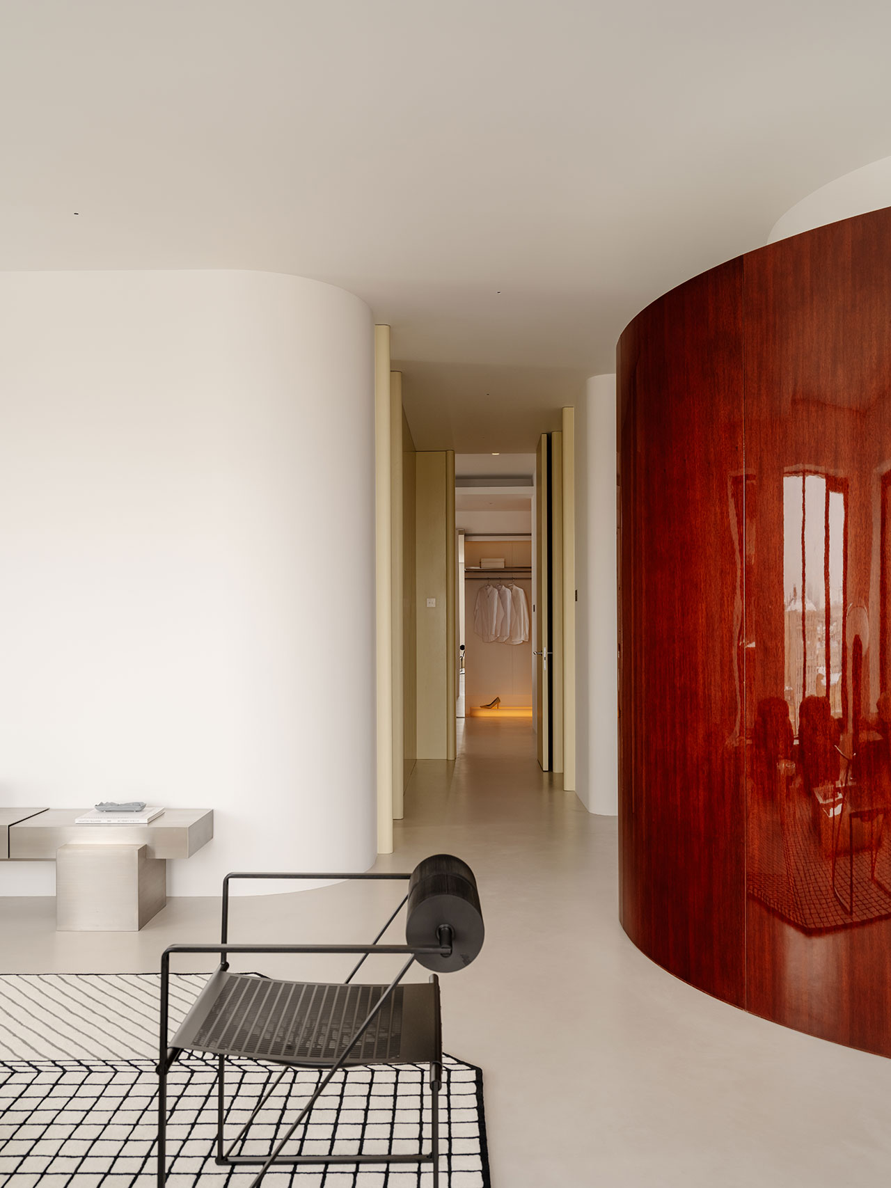 Minimalist interior with a black Antwe metal chair, a geometric rug, a low table, white curved walls, and a glossy curved wooden partition leading to a hallway with an open closet.