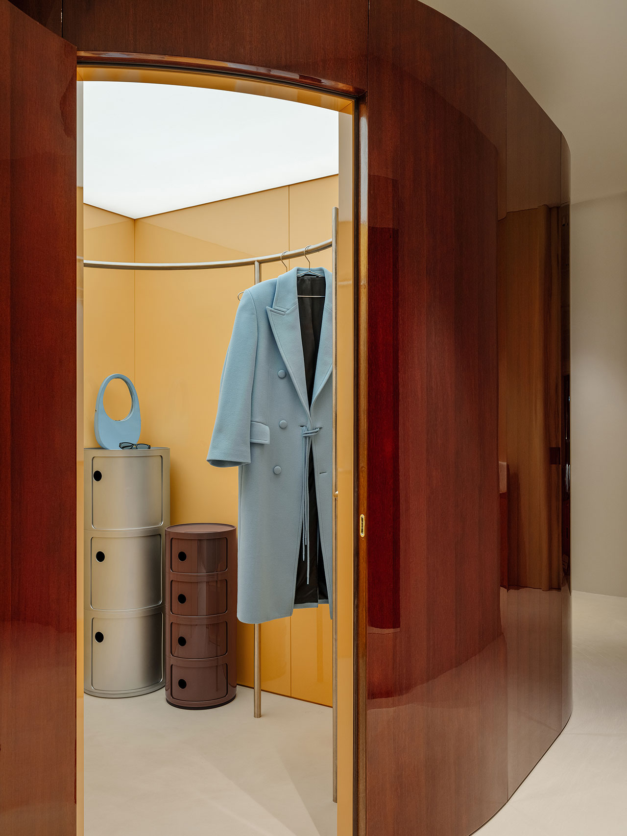 A modern Antwe closet with a yellow interior, displaying a light blue coat on a rack, a matching blue handbag, and three cylindrical storage units in silver and brown.