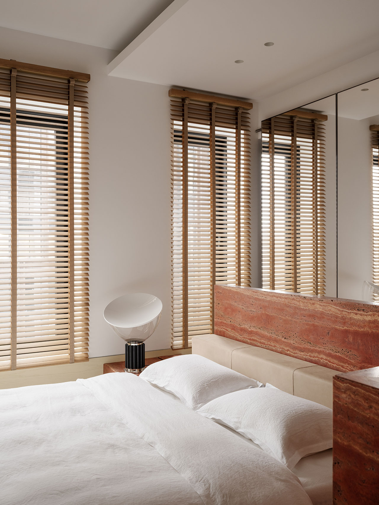 Minimalist Antwe bedroom with a white bed, travertine headboard, large mirrored wall, two tall windows with wooden blinds, and a modern white table lamp on a black stand.