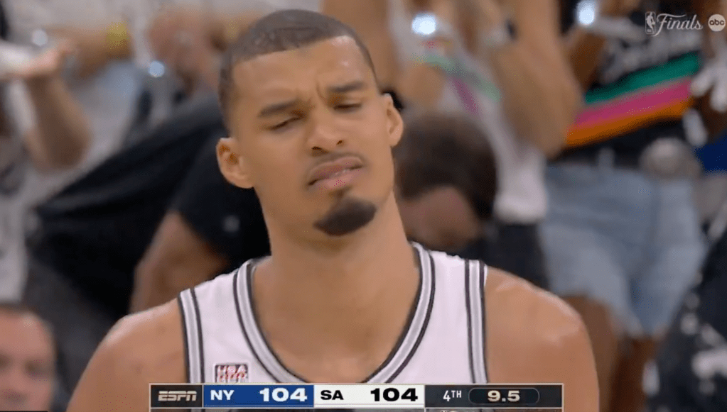 Basketball player with a beard and short hair looking up with a confused or displeased expression, wearing a white jersey with black and white trim.