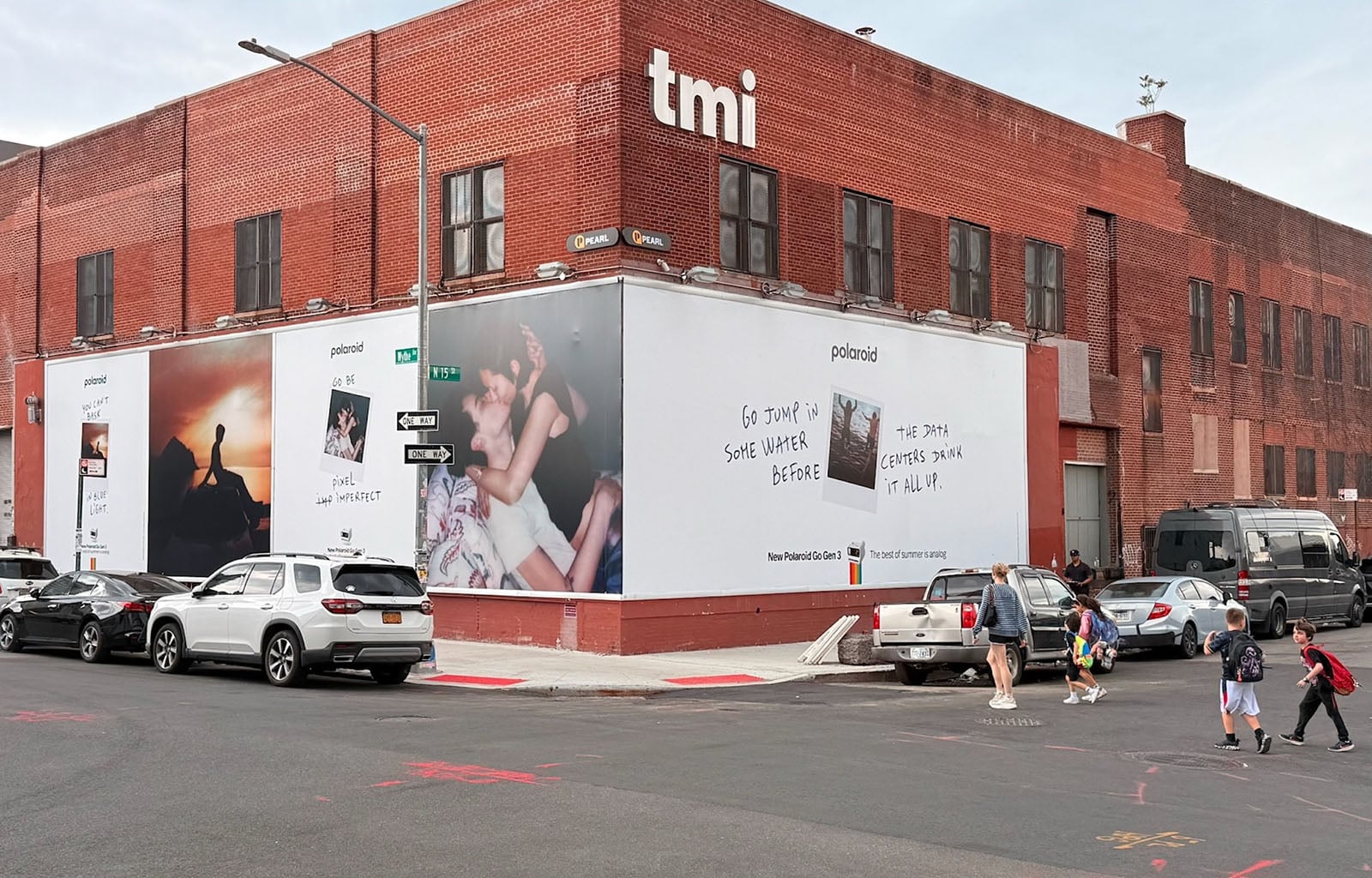 A brick building with large Polaroid ads on the side reading "Go jump in some water before the 29th graders show it all up." Several cars are parked nearby and people are walking on the sidewalk.