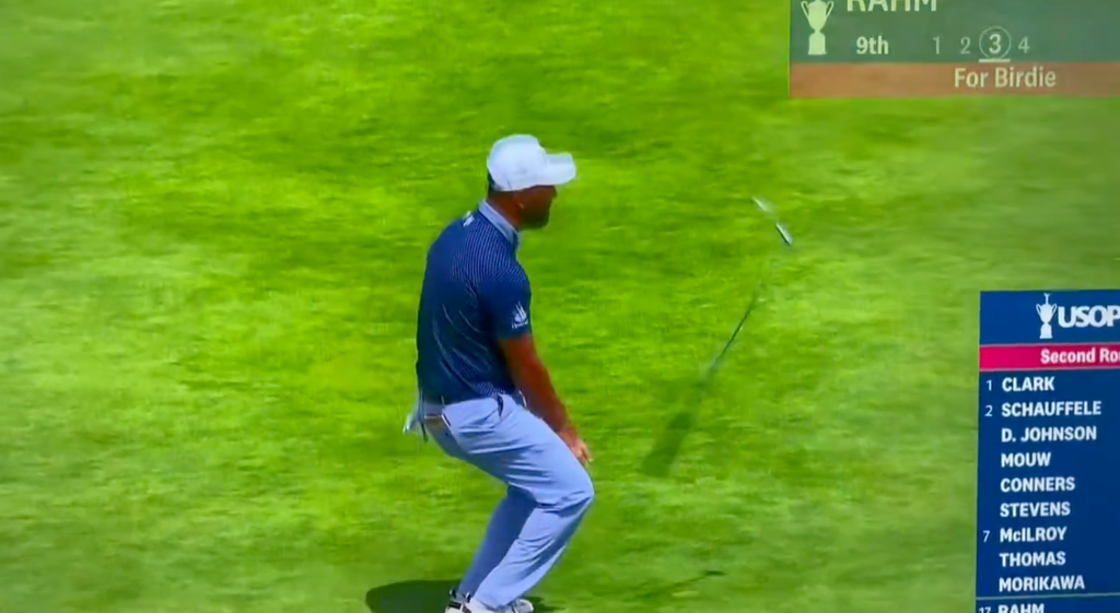 A male golfer in a white cap and blue shirt bends over, watching his golf club fly in the air after a shot, with a scoreboard visible on the right.