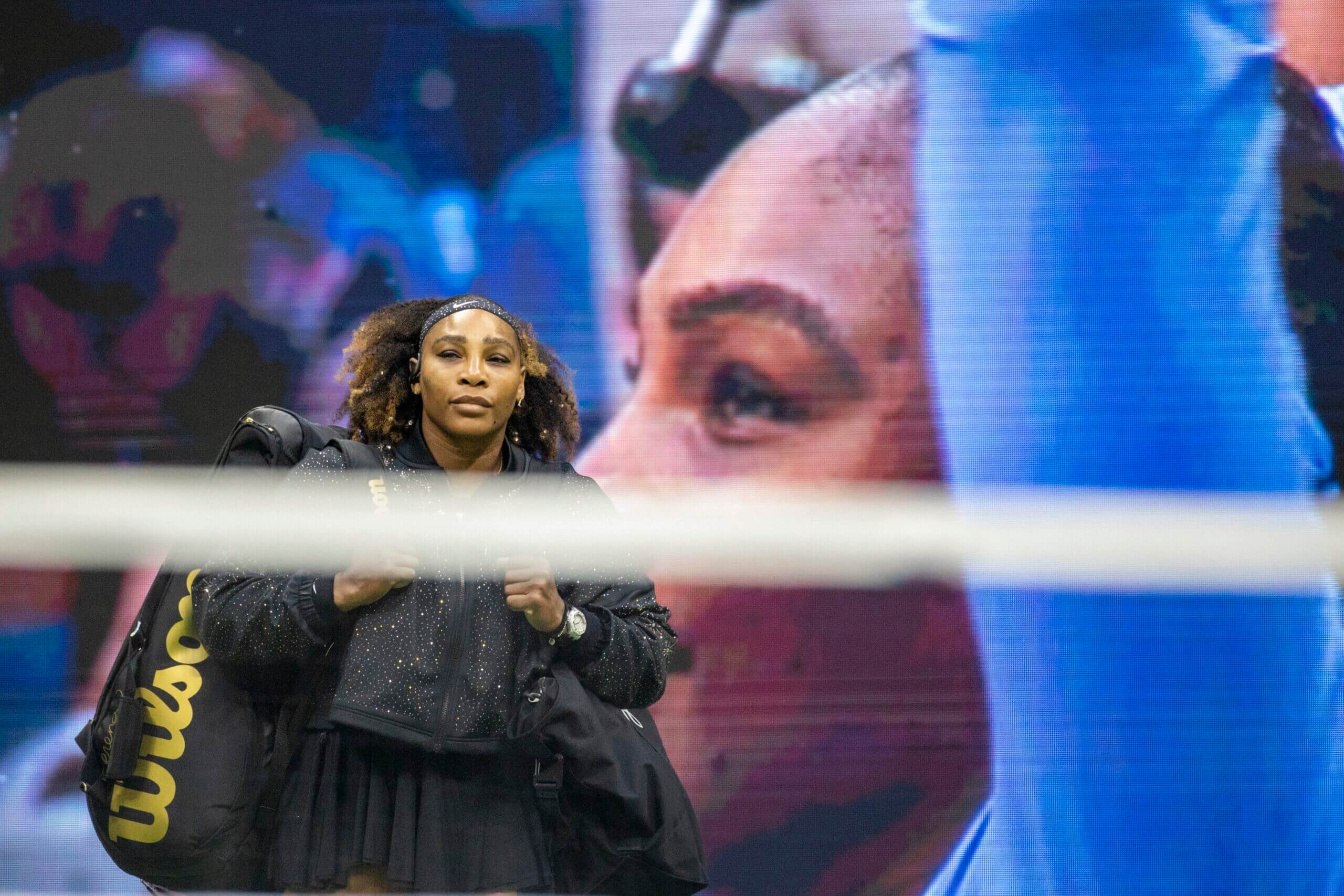 Serena Williams walks out onto a tennis court carrying a black-and-gold tennis bag, with a photo of herself celebrating on a big screen behind her.